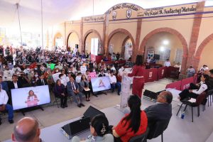 Conferencia o evento público con audiencia numerosa en un espacio cubierto con arcos de ladrillo y pantallas digitales.