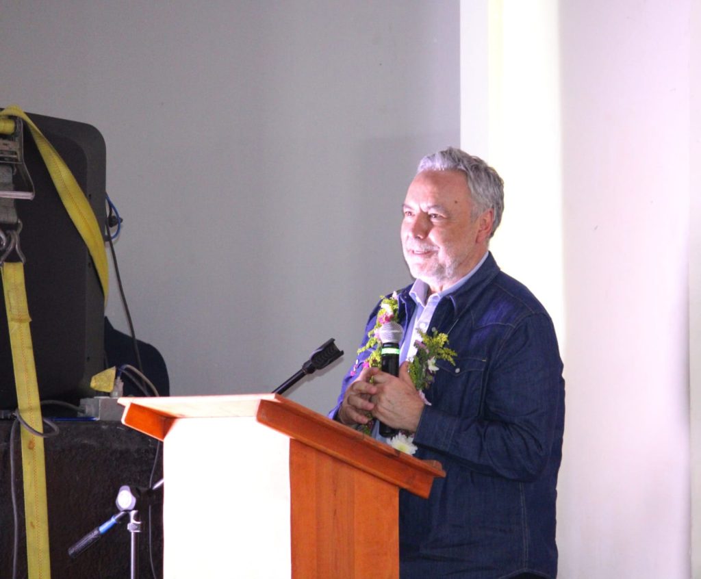 Hombre mayor con barba y cabello gris, vestido con chaqueta azul, hablando en un evento con un micrófono y decorado con flores.