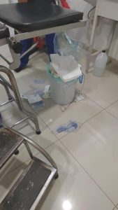 Ropa y objetos desordenados en un área de hospital, con una silla, un carrito y un cubo con papel y otros desechos en el suelo.