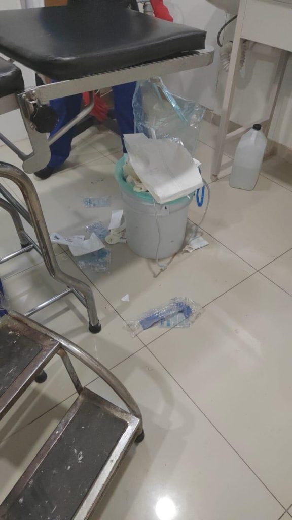 Ropa y objetos desordenados en un área de hospital, con una silla, un carrito y un cubo con papel y otros desechos en el suelo.
