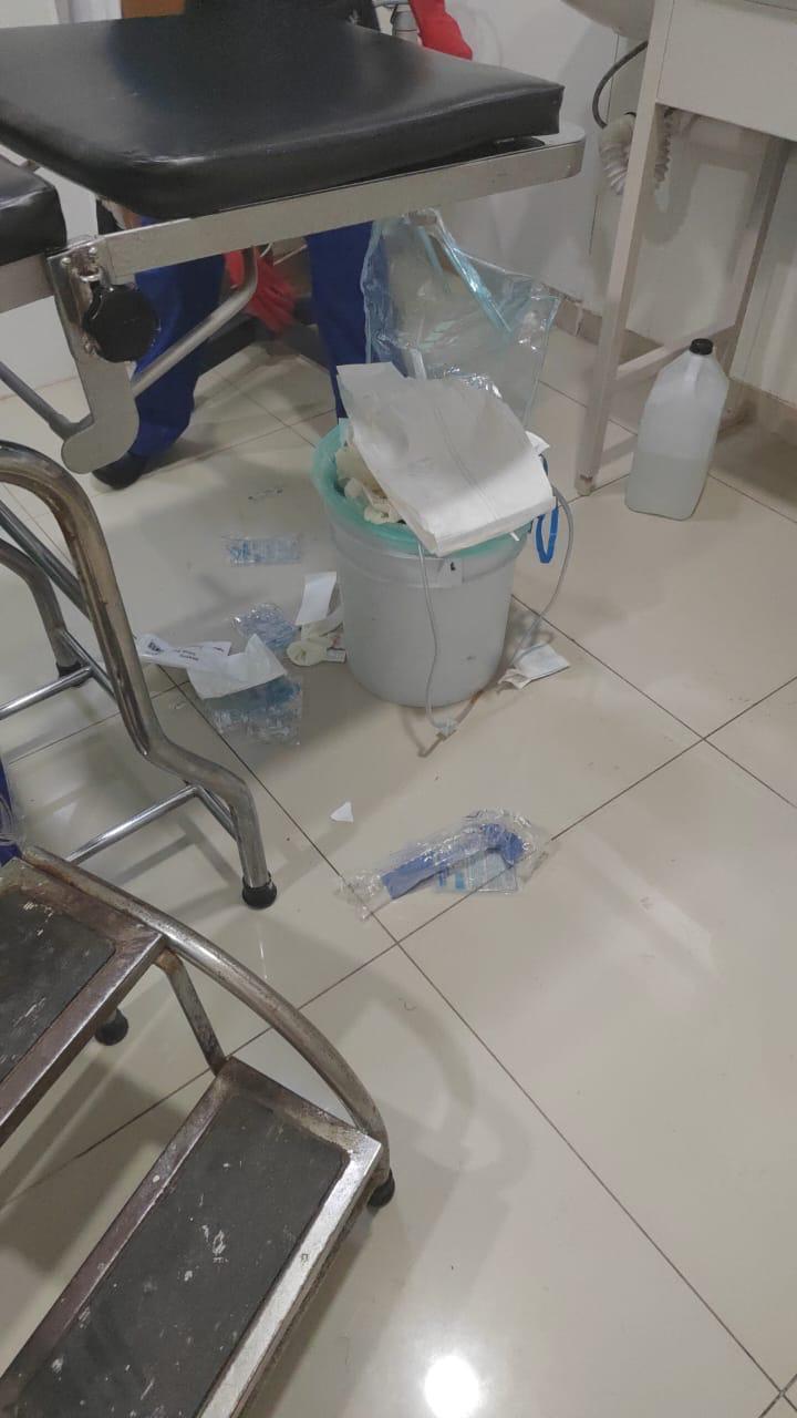 Ropa y objetos desordenados en un área de hospital, con una silla, un carrito y un cubo con papel y otros desechos en el suelo.
