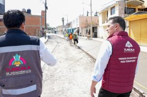Personal de Tlaxcala supervisando obras en una calle del municipio, con edificios y residentes en el fondo, en un día soleado y despejado.
