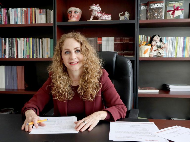 Mujer con cabello rizado y sonrisa, sentada en oficina con estanterías llenas de libros y decoraciones, en un entorno profesional y acogedor.