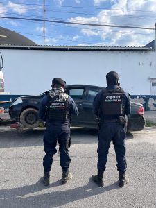Dos oficiales de Gopes Huamantla de espaldas, vigilando una calle con un coche dañado y un fondo de cielo con nubes y cables eléctricos.