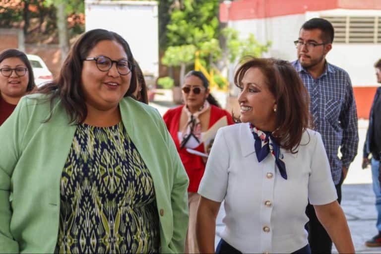 Amplia imagen de un grupo de personas conversando al aire libre, destacando a dos mujeres en primer plano, una con gafas y otra con blusa blanca, en un entorno urbano con árboles y edificios de fondo.