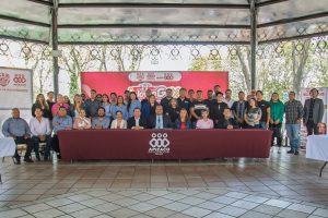 Grupo de personas en un evento oficial en un espacio abierto con estructura metálica y fondo con logotipos y pantallas digitales.