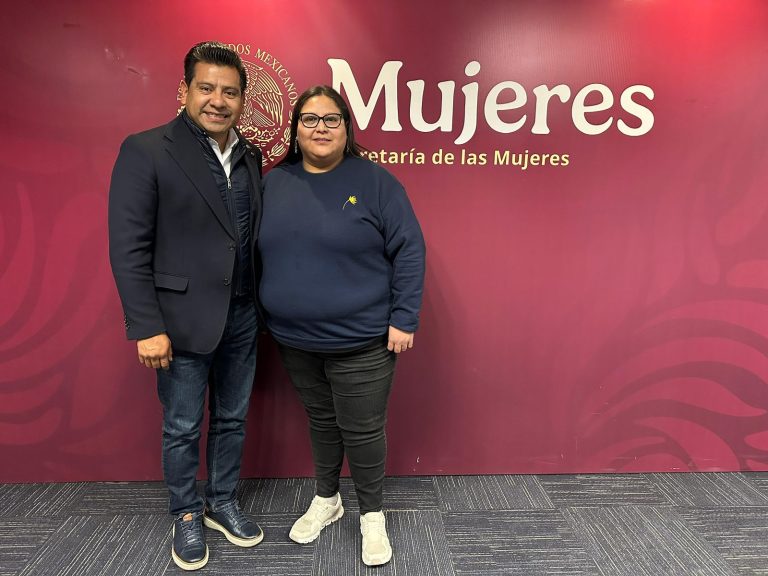 Personas en una oficina de la Secretaría de las Mujeres en Tlaxcala, posando para una foto en un evento oficial, con fondo en color rosa y logotipo institucional visible.