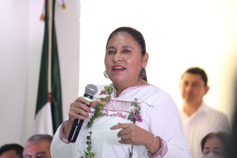 Una mujer en un evento público, hablando con un micrófono, vestida con ropa tradicional y adornada con flores, en un entorno formal con varias personas en el fondo.