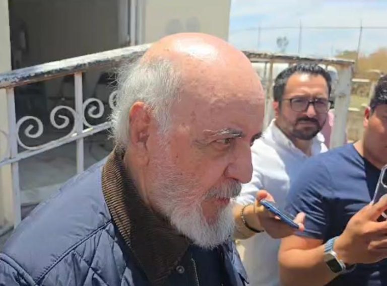 Un hombre mayor con barba blanca y cabello canoso, usando una chaqueta azul, en un entorno exterior, rodeado de varias personas que parecen periodistas o reporteros con teléfonos móviles y grabadoras.