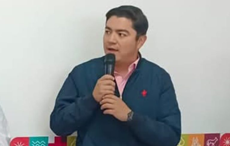 Hombre joven hablando en micrófono durante una conferencia o evento en interior, con fondo blanco y decoraciones coloridas en la parte inferior.