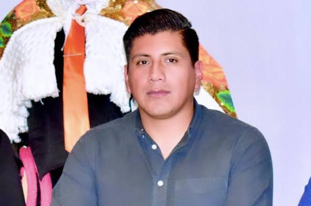 Hombre joven con cabello oscuro peinado hacia atrás, vestido con camisa azul y fondo de colores vibrantes.