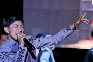 Un joven cantante con expresión emotiva mientras canta con un micrófono en mano, vestido con ropa tradicional mexicana en un escenario.