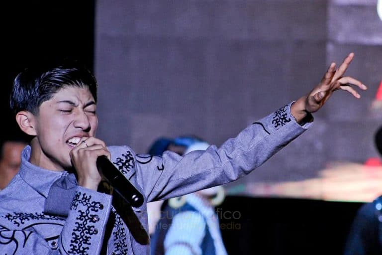 Un joven cantante con expresión emotiva mientras canta con un micrófono en mano, vestido con ropa tradicional mexicana en un escenario.
