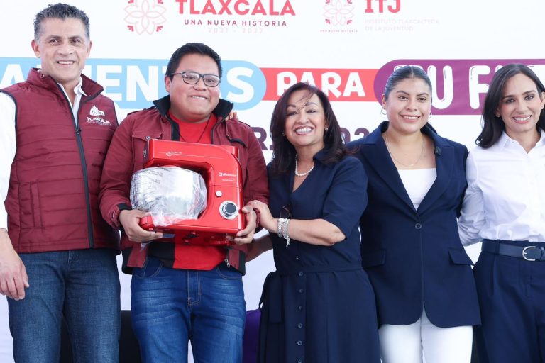 Cámara de personas en un evento, entregando un electrodoméstico a un joven, con un fondo de banner con logotipos y texto en español.