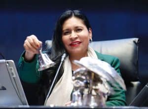 Mujer en una conferencia, sosteniendo un objeto metálico, con fondo oscuro y expresión sonriente, en un entorno formal y profesional.