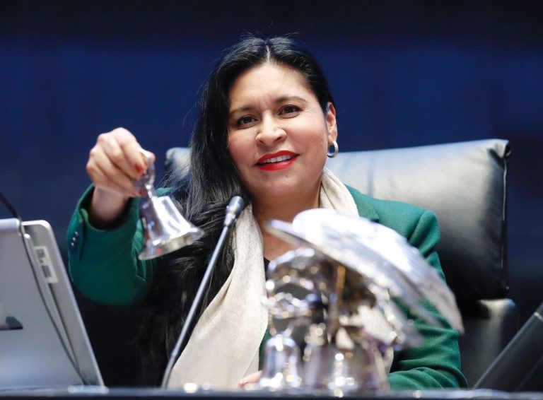 Mujer en una conferencia, sosteniendo un objeto metálico, con fondo oscuro y expresión sonriente, en un entorno formal y profesional.