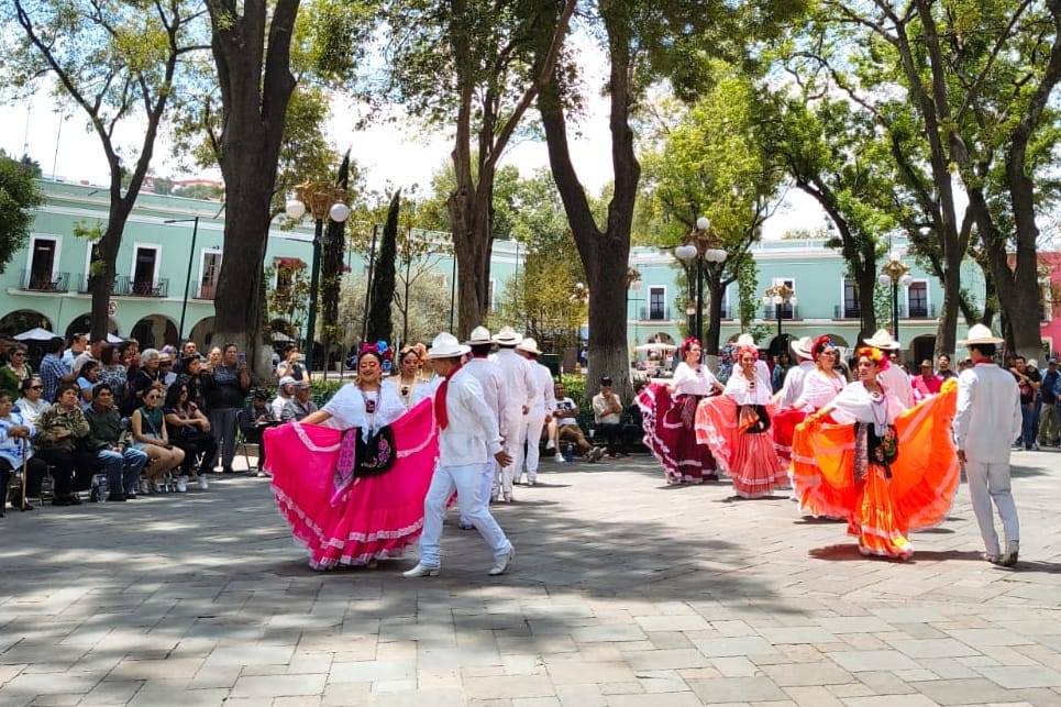 Grupo de personas bailando en una plaza pública rodeada de árboles y espectadores, con vestimenta tradicional mexicana y un ambiente festivo.