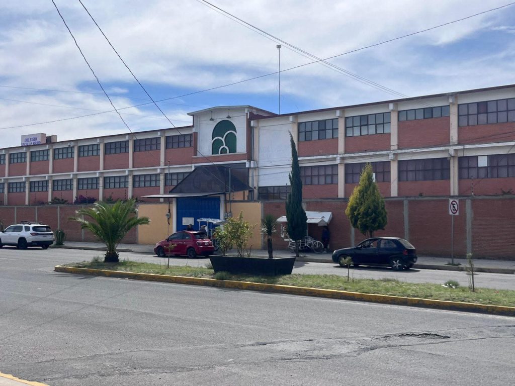 Árboles y plantas en la acera frente a un edificio de varios pisos en una calle de Tlaxcala, México, con vehículos estacionados y cables eléctricos en el cielo.