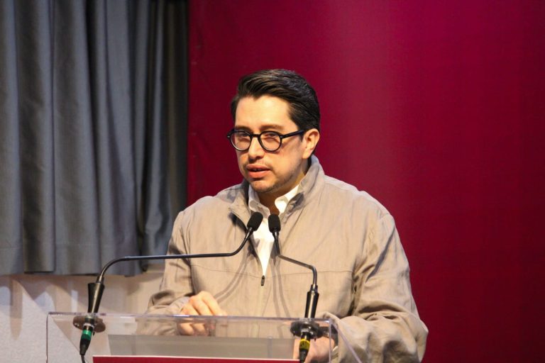 Hombre con gafas y chaqueta clara hablando en un podio durante una conferencia o evento formal.