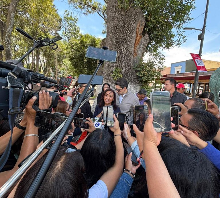Una mujer sonriente en medio de una multitud que la rodea, tomando fotos y videos con teléfonos móviles en un evento público al aire libre, con árboles y un edificio de fondo.