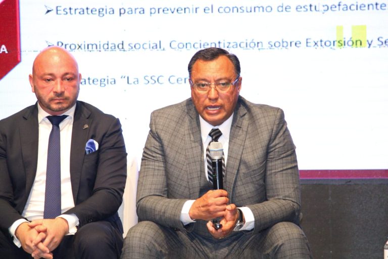 Dos hombres en una conferencia, uno con traje gris y gafas, hablando en un micrófono, mientras el otro escucha atentamente, en un entorno profesional con pantalla de fondo.