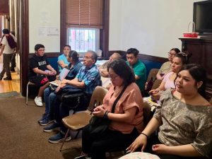 Grupo de personas sentadas en una sala de espera, algunas usando teléfonos móviles y otras conversando, en un ambiente interior con ventanas y paredes blancas.