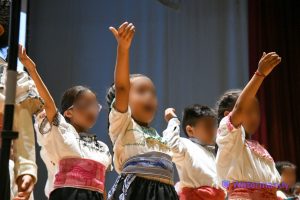Niños en una presentación cultural, vestidos con trajes tradicionales, en un escenario con cortinas rojas y fondo gris.