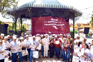 Trabajadores y participantes en una manifestación en un parque, sosteniendo carteles y certificados, con un escenario de fondo que tiene un banner en defensa de la transformación y la instalación de c.