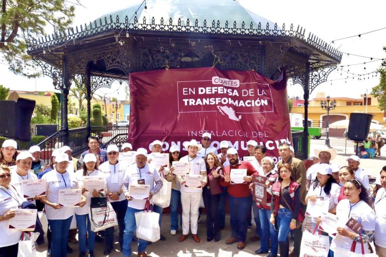 Trabajadores y participantes en una manifestación en un parque, sosteniendo carteles y certificados, con un escenario de fondo que tiene un banner en defensa de la transformación y la instalación de c.