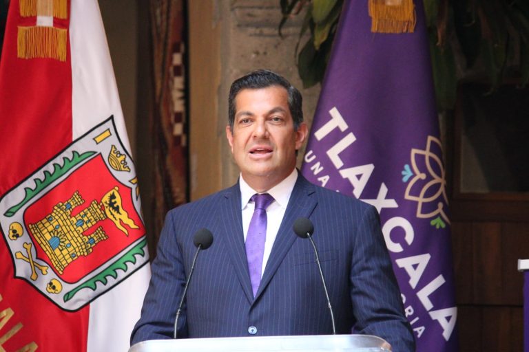 Hombre vestido con traje azul y corbata morada hablando en un podio con banderas de Tlaxcala y un logo de la ciudad en el fondo.