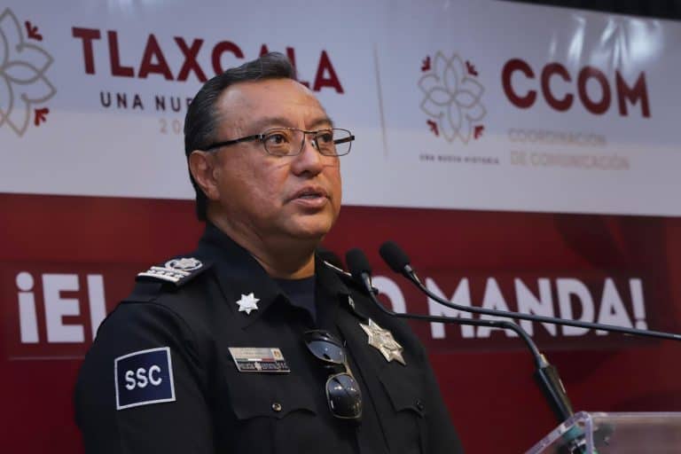 Policía de Tlaxcala en una conferencia de prensa, vistiendo uniforme y gafas, hablando frente a un micrófono en un evento oficial.