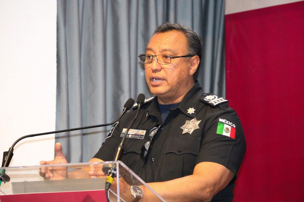 Policía de México en uniforme, hablando en una conferencia de prensa, con fondo de cortinas grises y bandera de México en el escenario.