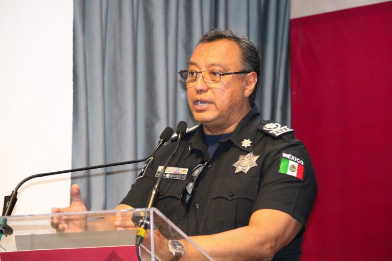 Policía de México en uniforme, hablando en una conferencia de prensa, con fondo de cortinas grises y bandera de México en el escenario.