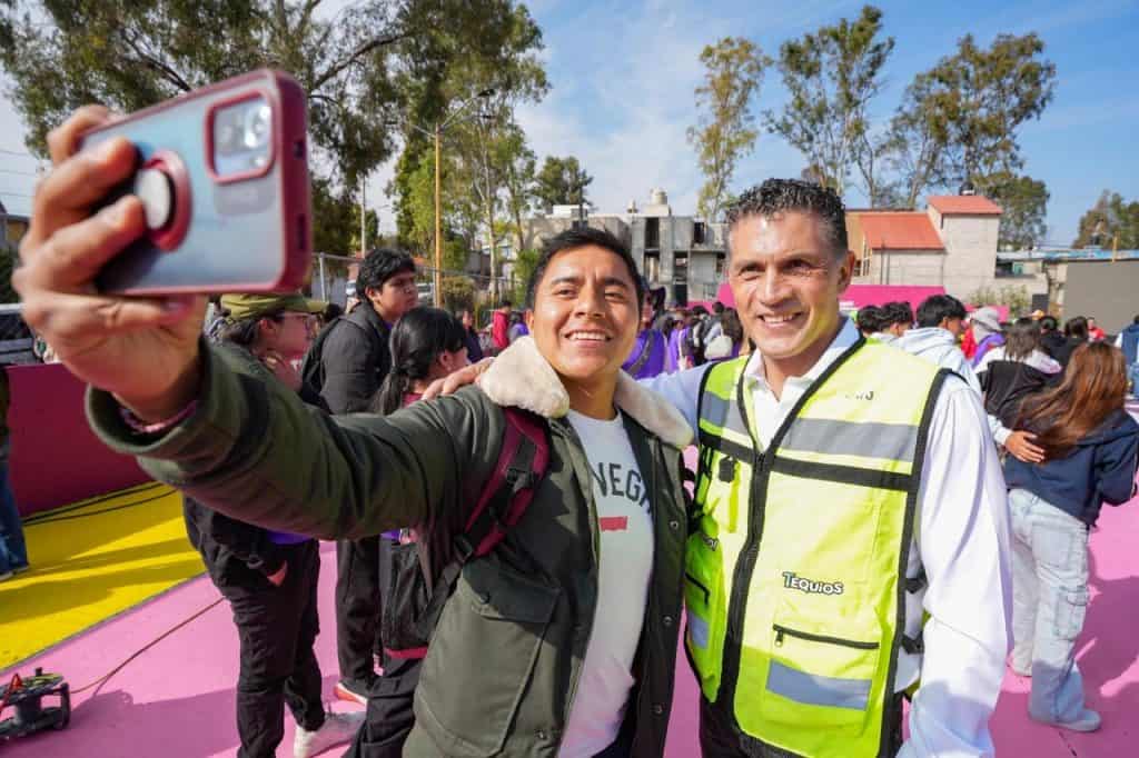 Tomando una selfie en un evento público con muchas personas en el fondo, en un día soleado y con árboles, mostrando alegría y convivencia comunitaria.