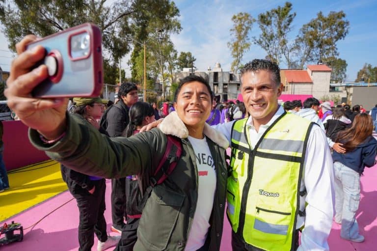 Tomando una selfie en un evento público con muchas personas en el fondo, en un día soleado y con árboles, mostrando alegría y convivencia comunitaria.