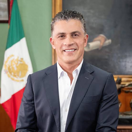 Hombre en traje formal con bandera de México al fondo, en un entorno oficial y profesional.