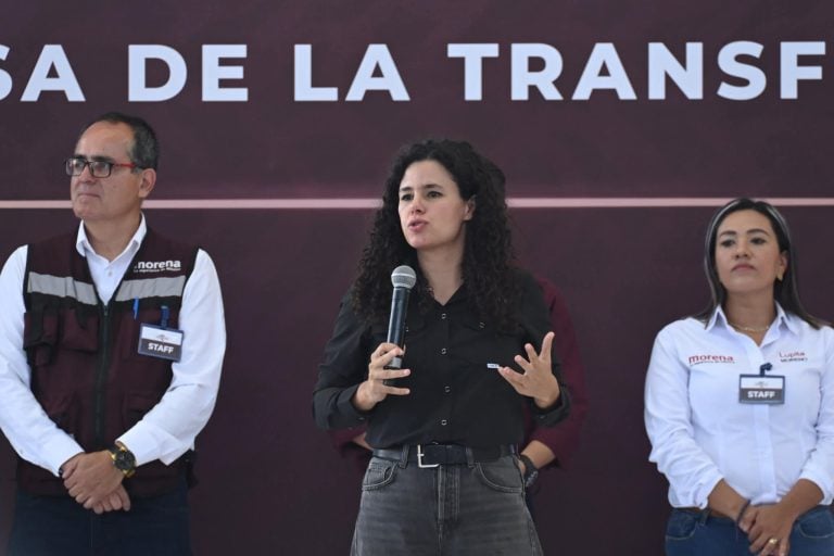 Asistente de la imagen en la conferencia de prensa con tres personas en el escenario, una mujer en el centro hablando con micrófono y dos personas a su lado, fondo con texto en español.