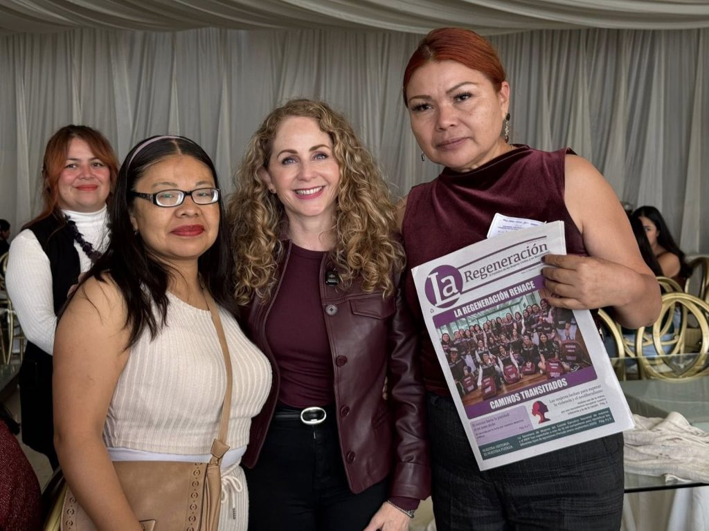 Grupo de mujeres en evento, una de ellas sostiene un periódico con el título "Regeneración" en un entorno interior con cortinas blancas.