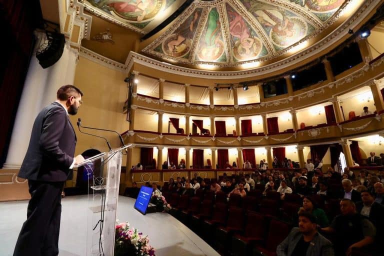 Conferencista en un auditorio con techo decorado y público asistente en una conferencia o evento formal.