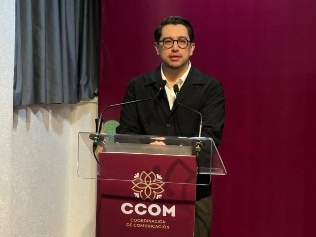 El hombre con gafas y chaqueta negra hablando en un podio con logotipo de CCOM en un evento formal.