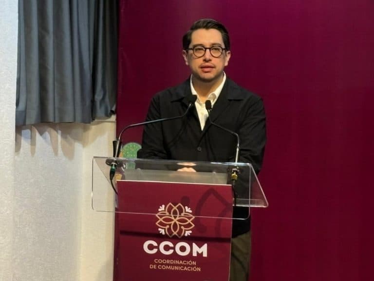 El hombre con gafas y chaqueta negra hablando en un podio con logotipo de CCOM en un evento formal.