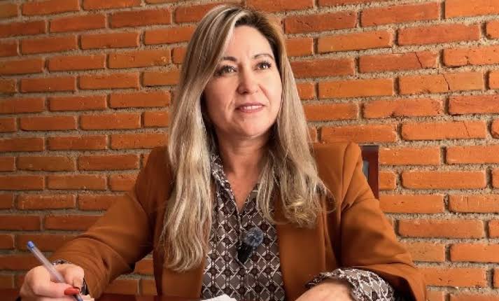Mujer sentada en una mesa con fondo de pared de ladrillos, sosteniendo un bolígrafo y sonriendo, en un entorno de oficina o entrevista.