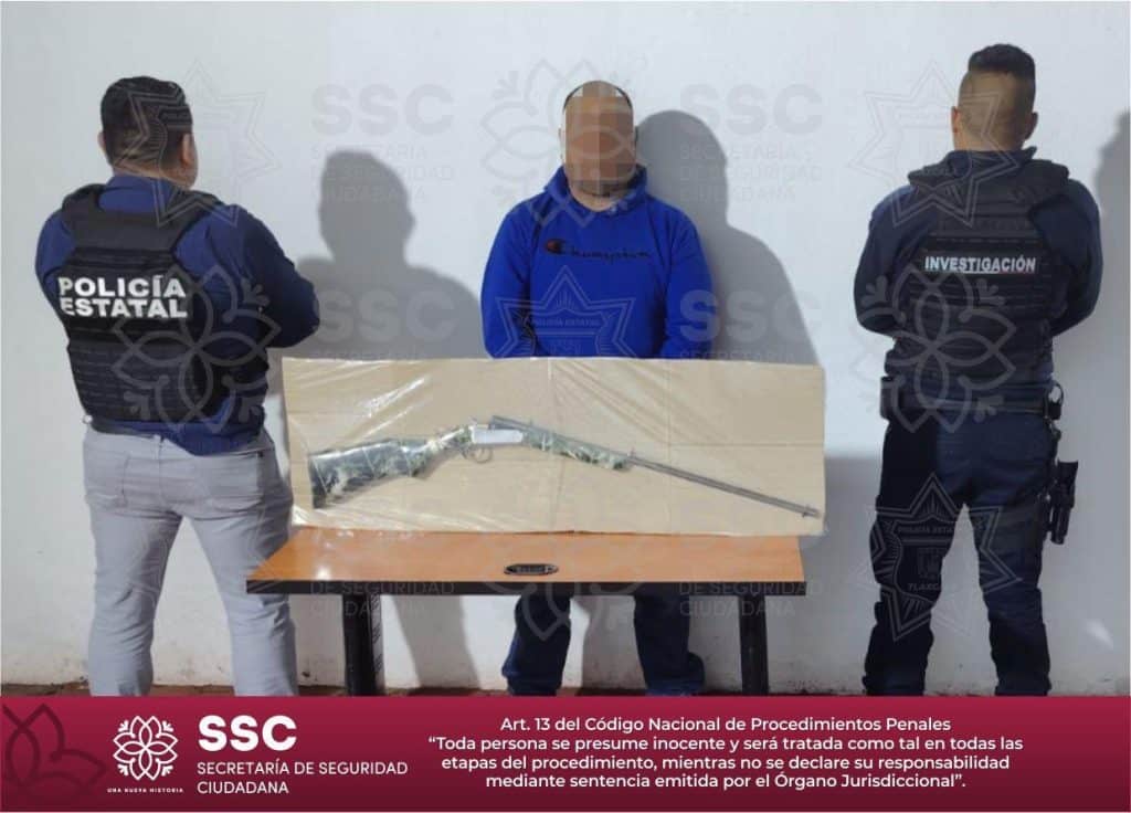 Persona detenido entre dos policías estatales, con un arma asegurada sobre una mesa, en una escena de investigación policial en Tlaxcala, México.