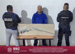 Persona detenido entre dos policías estatales, con un arma asegurada sobre una mesa, en una escena de investigación policial en Tlaxcala, México.