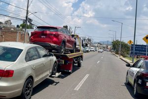 Remolque de autos en una calle de Tlaxcala, México, con varios vehículos en fila y un coche rojo en la plataforma del remolque, en un día soleado con cielo parcialmente nublado.