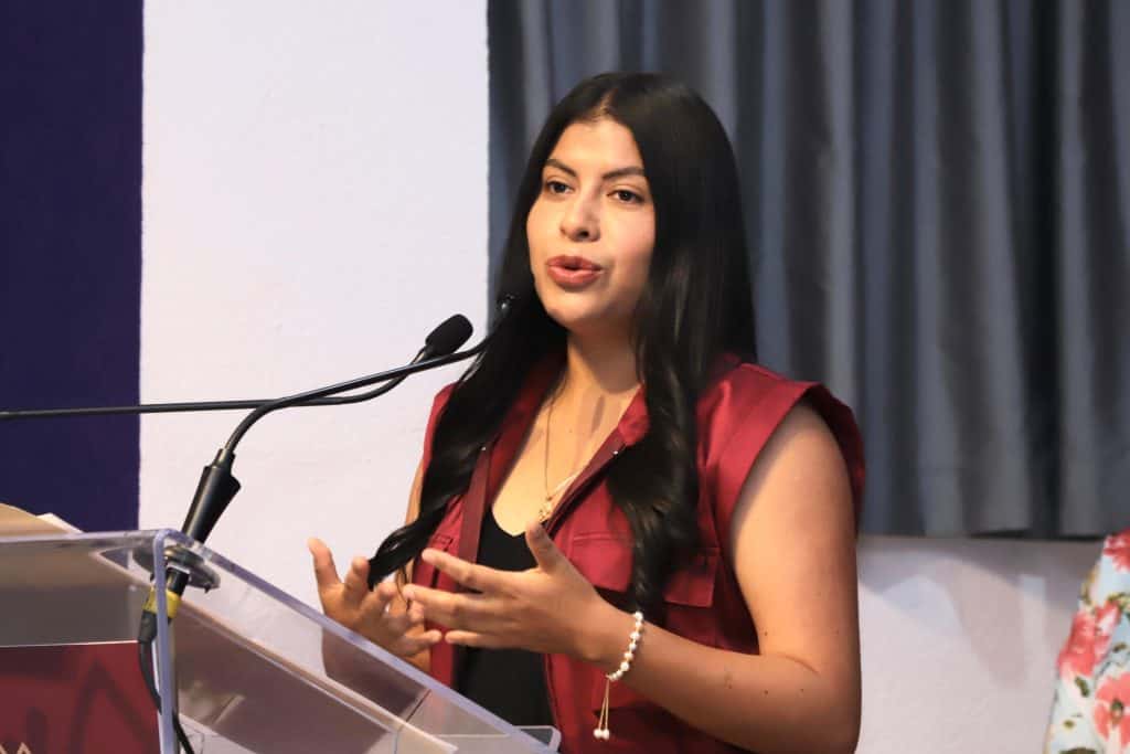 Mujer hablando en conferencia, vestida con blusa roja y cabello largo, en un evento formal con micrófono y fondo de cortinas grises.