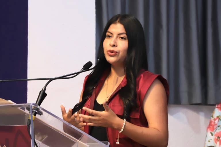 Mujer hablando en conferencia, vestida con blusa roja y cabello largo, en un evento formal con micrófono y fondo de cortinas grises.