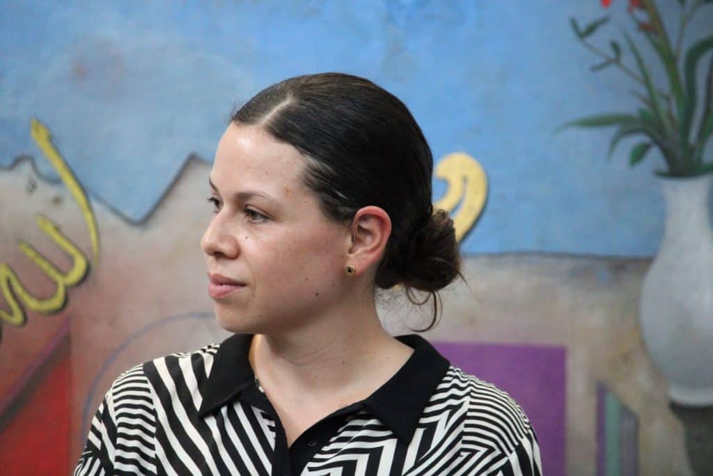 Mujer con cabello recogido y blusa a rayas en un entorno artístico con mural colorido de fondo.