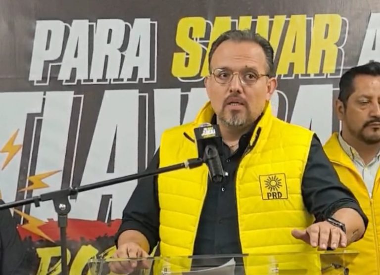 El hombre con gafas y barba, vestido con un chaleco amarillo con el logo del PRD, habla en una conferencia de prensa con un micrófono frente a un cartel de fondo.