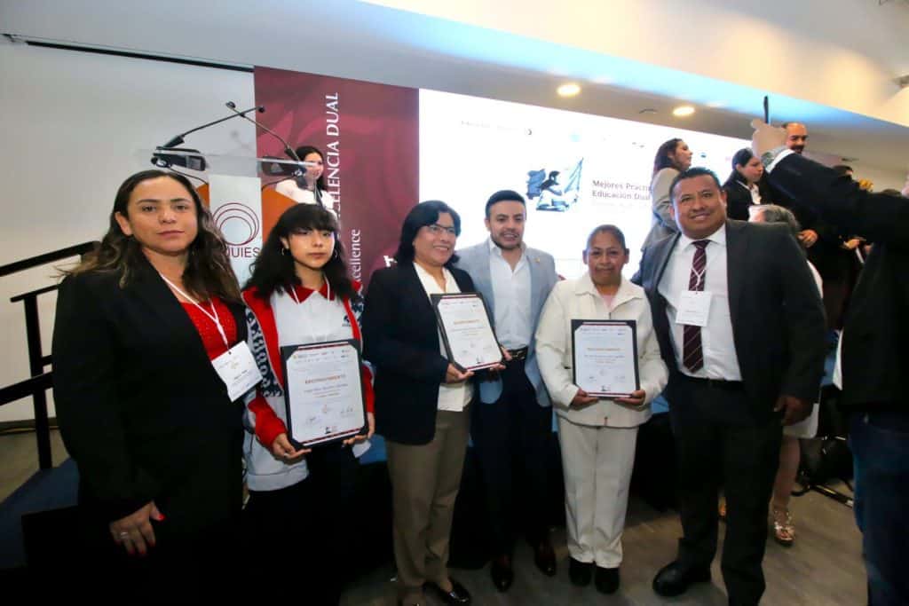 Grupo de personas en una ceremonia de reconocimiento, algunos sosteniendo certificados, en un evento formal en un auditorio con pantalla grande al fondo.